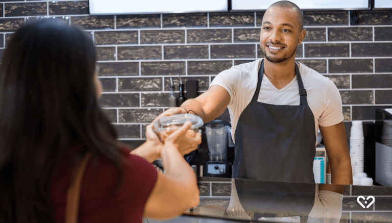Lächelnder Mann mit Schürze überreicht einer Kundin einen Essensbehälter an der Take-away-Theke.