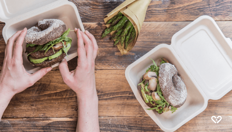 Hände halten Gourmet-Burger in umweltfreundlicher Take-away-Box, zweiter Burger und Spargel auf Holztisch.