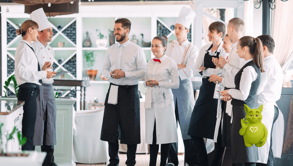Besprechung vom Restaurantteam in Uniform, Tuutuu-Maskottchen zeigt OK-Geste am Rand.