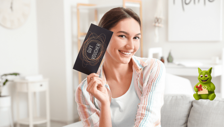 Lächelnde Frau hält einen schwarzen Geschenkgutschein; Tuutuu mit Umschlag sitzt in der Ecke.